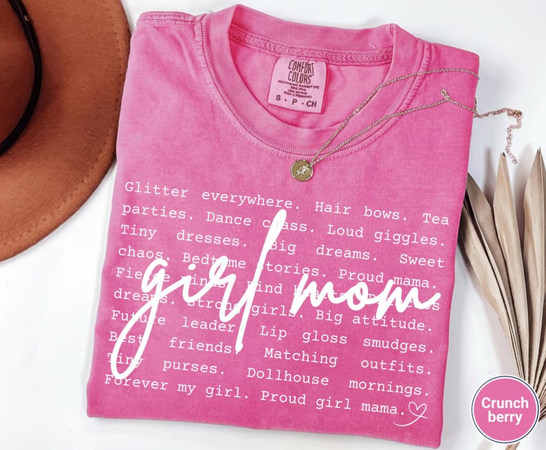 May include: A pink Comfort Colors t-shirt with the words "girl mom" in white script. The shirt features a list of related words like "glitter," "dreams," and "proud mama." A gold necklace with a pendant is draped on the shirt.