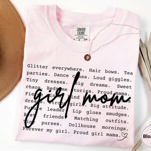 May include: A pale pink Comfort Colors t-shirt with the words "girl mom" in a handwritten font. The shirt is covered in words like "glitter," "dreams," and "matching outfits." A gold necklace and a dried plant are also visible.