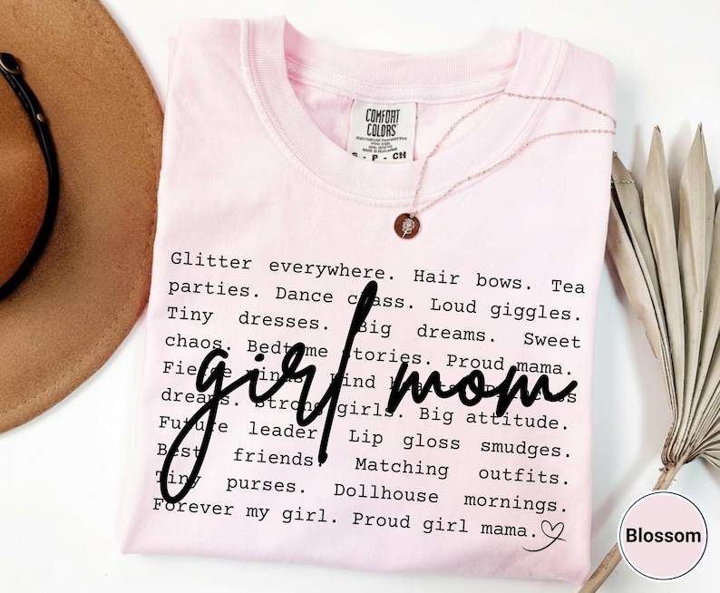 May include: A pale pink Comfort Colors t-shirt with the words "girl mom" in a handwritten font. The shirt is covered in words like "glitter," "dreams," and "matching outfits." A gold necklace and a dried plant are also visible.