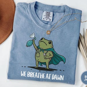 Pode incluir: Uma t-shirt azul jeans com o gr&aacute;fico de um sapo verde vestindo uma capa azul-petr&oacute;leo e um estetosc&oacute;pio, segurando um bal&atilde;o de reanima&ccedil;&atilde;o. O texto "WE BREATHE AT DAWN" est&aacute; impresso em branco abaixo do sapo. Etiqueta Comfort Colors vis&iacute;vel.