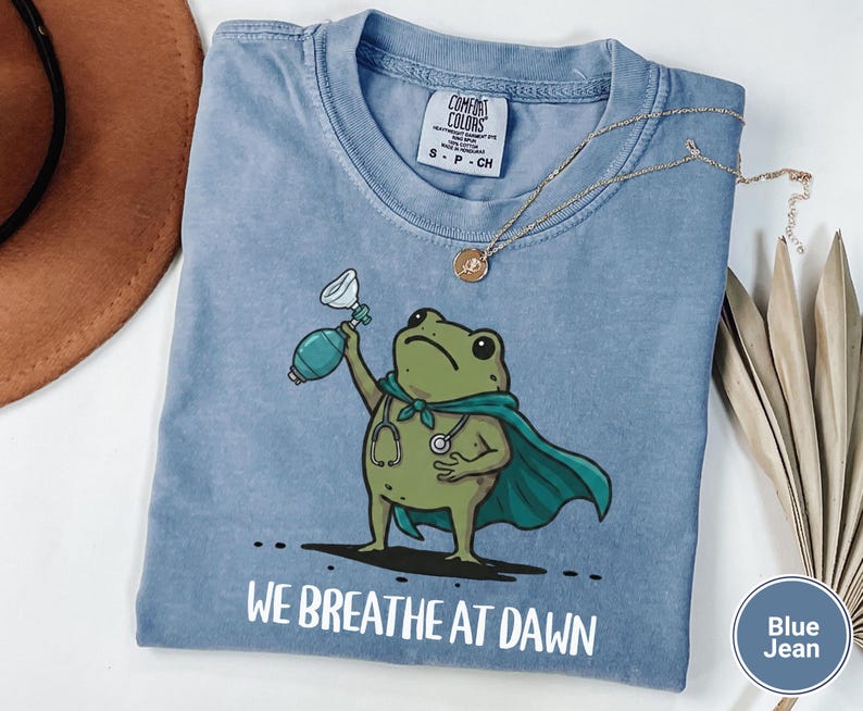 Pode incluir: Uma t-shirt azul jeans com o gr&aacute;fico de um sapo verde vestindo uma capa azul-petr&oacute;leo e um estetosc&oacute;pio, segurando um bal&atilde;o de reanima&ccedil;&atilde;o. O texto "WE BREATHE AT DAWN" est&aacute; impresso em branco abaixo do sapo. Etiqueta Comfort Colors vis&iacute;vel.