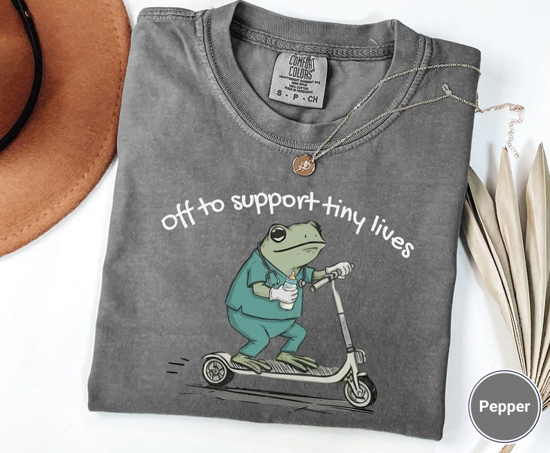 May include: Pepper-colored t-shirt with a frog graphic riding a scooter, wearing scrubs and holding a bottle. The text "off to support tiny lives" is above the frog. A silver necklace with a pendant is on the shirt.