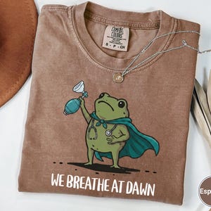 Pode incluir: Uma t-shirt cor de caf&eacute; expresso com um gr&aacute;fico de um sapo verde a usar uma capa azul-petr&oacute;leo e um estetosc&oacute;pio, segurando um ressuscitador manual. Abaixo do sapo, o texto diz WE BREATHE AT DAWN em letras mai&uacute;sculas brancas.