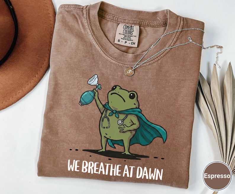 Pode incluir: Uma t-shirt cor de caf&eacute; expresso com um gr&aacute;fico de um sapo verde a usar uma capa azul-petr&oacute;leo e um estetosc&oacute;pio, segurando um ressuscitador manual. Abaixo do sapo, o texto diz WE BREATHE AT DAWN em letras mai&uacute;sculas brancas.