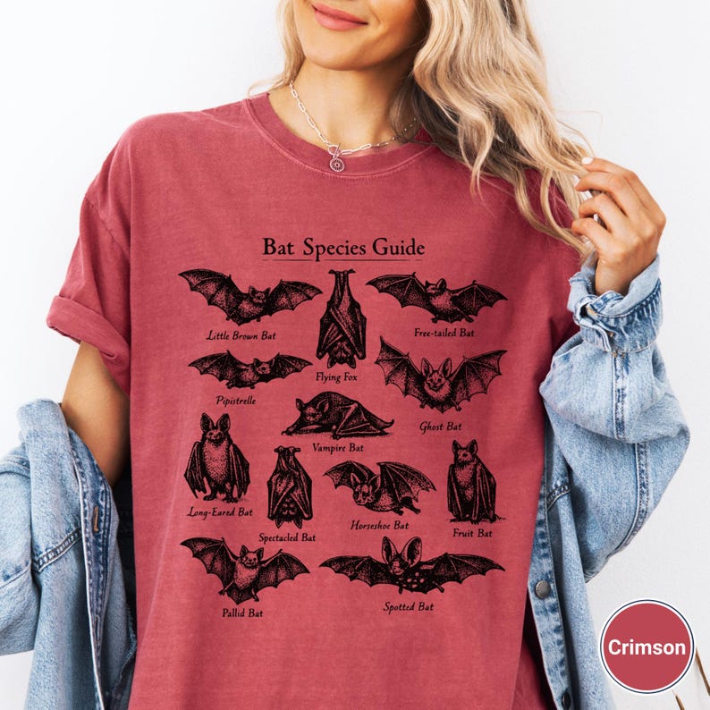 May include: Crimson-colored t-shirt featuring a "Bat Species Guide" graphic. The graphic includes illustrations of various bat species, such as the Little Brown Bat, Flying Fox, and Vampire Bat. The shirt is made of a soft material.