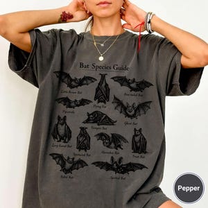 May include: Dark gray t-shirt featuring a "Bat Species Guide" with various bat illustrations. The shirt includes the names of different bat species. The shirt is a relaxed fit.