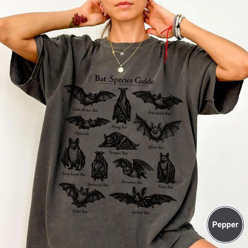 May include: Dark gray t-shirt featuring a "Bat Species Guide" with various bat illustrations. The shirt includes the names of different bat species. The shirt is a relaxed fit.