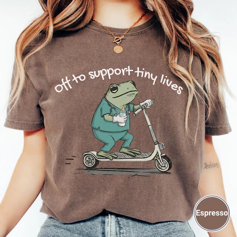 May include: Espresso-colored t-shirt featuring a cartoon frog in scrubs riding a scooter, holding a baby bottle. The text "off to support tiny lives" is arched above the frog. The shirt is a casual style.