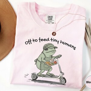 May include: Light pink Comfort Colors t-shirt with the phrase "off to feed tiny humans" and a cartoon frog riding a scooter while carrying a tray of food. The frog wears a hat and apron. A necklace is draped across the shirt.