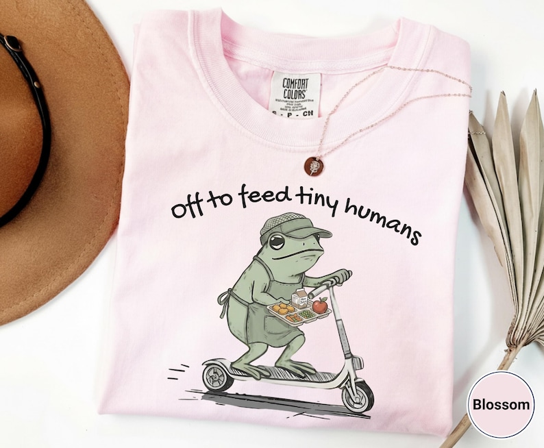May include: Light pink Comfort Colors t-shirt with the phrase "off to feed tiny humans" and a cartoon frog riding a scooter while carrying a tray of food. The frog wears a hat and apron. A necklace is draped across the shirt.