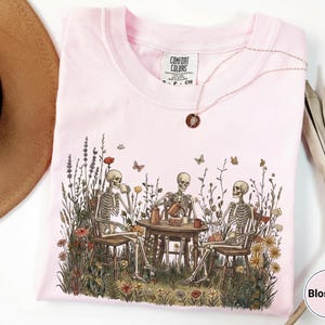Może przedstawiać: Jasnor&oacute;żowy t-shirt z grafiką przedstawiającą trzy szkielety siedzące przy okrągłym stole na polu pełnym polnych kwiat&oacute;w i pijące z kubk&oacute;w. Koszulka posiada metkę marki Comfort Colors przy szyi oraz mały naszyjnik z okrągłą zawieszką.