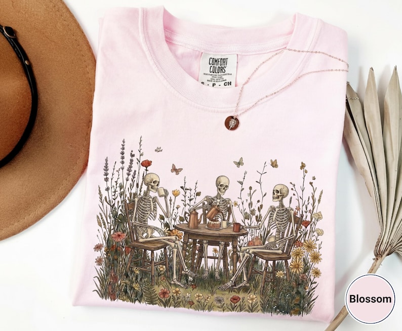Może przedstawiać: Jasnor&oacute;żowy t-shirt z grafiką przedstawiającą trzy szkielety siedzące przy okrągłym stole na polu pełnym polnych kwiat&oacute;w i pijące z kubk&oacute;w. Koszulka posiada metkę marki Comfort Colors przy szyi oraz mały naszyjnik z okrągłą zawieszką.