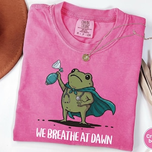 Pode incluir: Uma t-shirt de manga curta rosa brilhante com um gr&aacute;fico de um sapo verde usando uma capa azul-petr&oacute;leo e um estetosc&oacute;pio. O sapo segura um bal&atilde;o de reanima&ccedil;&atilde;o azul-petr&oacute;leo. Abaixo, o texto diz WE BREATHE AT DAWN em letras brancas.