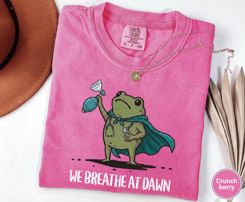 Pode incluir: Uma t-shirt de manga curta rosa brilhante com um gr&aacute;fico de um sapo verde usando uma capa azul-petr&oacute;leo e um estetosc&oacute;pio. O sapo segura um bal&atilde;o de reanima&ccedil;&atilde;o azul-petr&oacute;leo. Abaixo, o texto diz WE BREATHE AT DAWN em letras brancas.