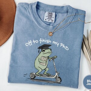 May include: Blue jean Comfort Colors t-shirt with the text "off to finish my PhD." The shirt features a cartoon frog wearing a graduation cap and riding a scooter. The frog is holding a stack of books labeled "Draft, Research, Economics."