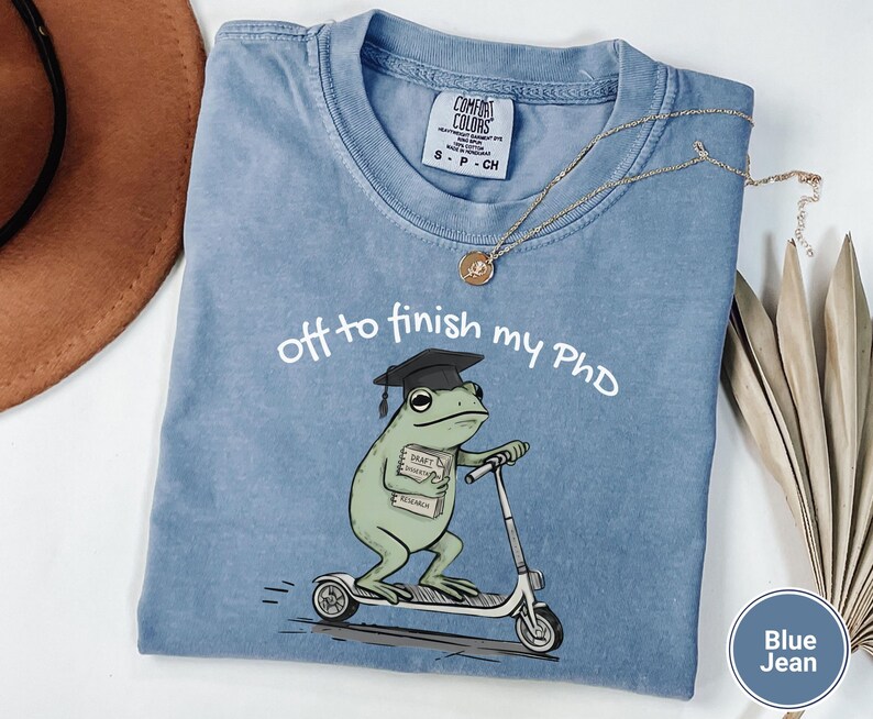 May include: Blue jean Comfort Colors t-shirt with the text "off to finish my PhD." The shirt features a cartoon frog wearing a graduation cap and riding a scooter. The frog is holding a stack of books labeled "Draft, Research, Economics."