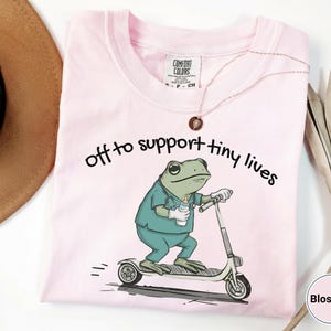 May include: Light pink t-shirt with the text "off to support tiny lives." The shirt features a cartoon frog in scrubs riding a scooter and holding a baby bottle. The shirt is a Comfort Colors brand.