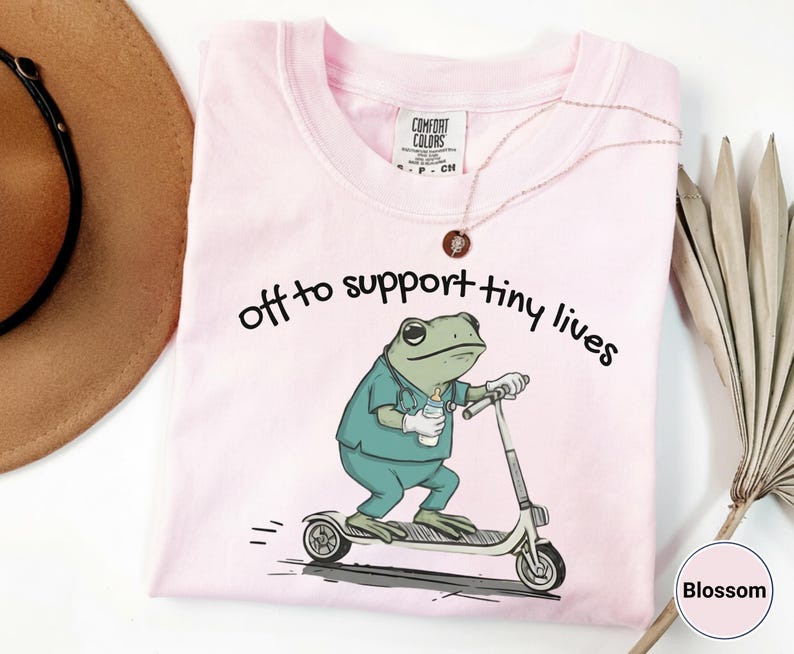 May include: Light pink t-shirt with the text "off to support tiny lives." The shirt features a cartoon frog in scrubs riding a scooter and holding a baby bottle. The shirt is a Comfort Colors brand.