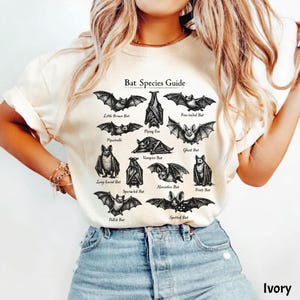 May include: Ivory-colored t-shirt featuring a detailed illustration of various bat species, labeled with their common names. The graphic includes bats like the Little Brown Bat, Flying Fox, and Vampire Bat. The text "Bat Species Guide" is at the top.