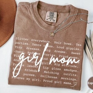 May include: Espresso-coloured t-shirt with the words "girl mom" in a cursive font. The shirt features a list of words like "glitter," "parties," and "big dreams." A silver necklace with a small pendant rests on the shirt.