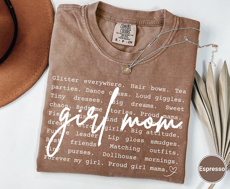 May include: Espresso-coloured t-shirt with the words "girl mom" in a cursive font. The shirt features a list of words like "glitter," "parties," and "big dreams." A silver necklace with a small pendant rests on the shirt.