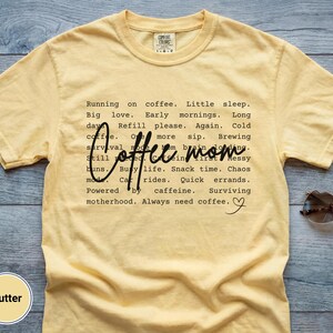 May include: Butter-colored t-shirt with the words "Coffee mom" in a script font. The shirt has a list of coffee-related phrases in a black, sans-serif font. Sunglasses rest on the shirt.