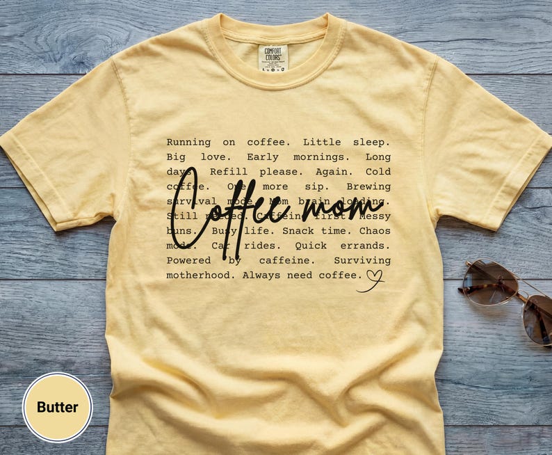 May include: Butter-colored t-shirt with the words "Coffee mom" in a script font. The shirt has a list of coffee-related phrases in a black, sans-serif font. Sunglasses rest on the shirt.