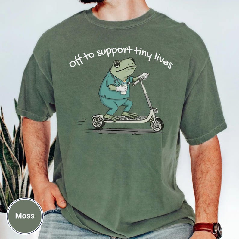 May include: Moss green t-shirt featuring a cartoon frog wearing scrubs and riding a scooter, holding a coffee cup and a baby bottle. The text "off to support tiny lives" is arched above the frog.