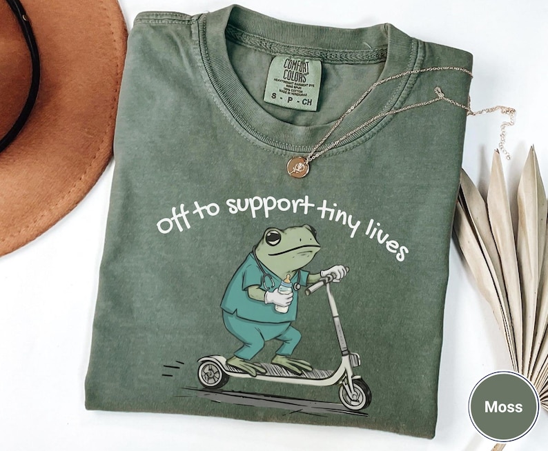 May include: Moss green t-shirt with the phrase "off to support tiny lives" and a cartoon frog riding a scooter. The frog is wearing scrubs and holding a baby bottle. A gold necklace is draped across the shirt.