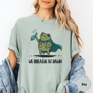 Pode incluir: Uma t-shirt verde s&aacute;lvia com um gr&aacute;fico de um sapo verde a usar uma capa azul-petr&oacute;leo e um estetosc&oacute;pio ao pesco&ccedil;o. O sapo segura um bal&atilde;o de reanima&ccedil;&atilde;o azul-petr&oacute;leo. Abaixo, o texto diz WE BREATHE AT DAWN em letras pretas.