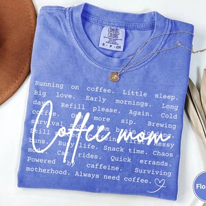 May include: A blue Comfort Colors t-shirt with the words "Coffee mom" in white script. The shirt has a list of phrases about motherhood and coffee. A gold necklace with a pendant is on the shirt.