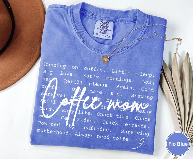 May include: A blue Comfort Colors t-shirt with the words "Coffee mom" in white script. The shirt has a list of phrases about motherhood and coffee. A gold necklace with a pendant is on the shirt.