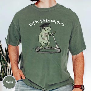May include: Moss green t-shirt with the text "off to finish my PhD". The graphic shows a frog wearing a graduation cap and riding a scooter, holding a book labeled "Draft, Research".
