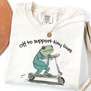 May include: Cream-colored t-shirt with the text "off to support tiny lives." The shirt features a cartoon frog wearing scrubs and riding a scooter while holding a baby bottle. A gold necklace and a brown hat are visible.