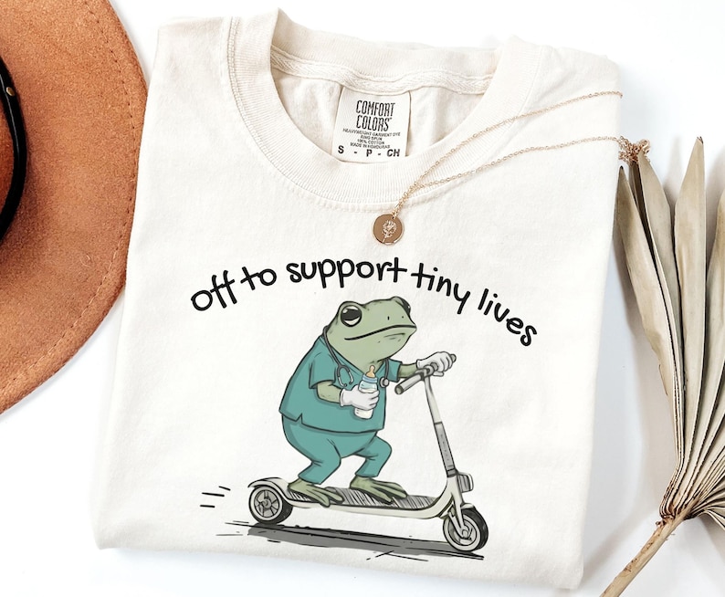 May include: Cream-colored t-shirt with the text "off to support tiny lives." The shirt features a cartoon frog wearing scrubs and riding a scooter while holding a baby bottle. A gold necklace and a brown hat are visible.