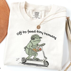 May include: Ivory-colored t-shirt with the phrase "off to feed tiny humans" in black script. The shirt features a cartoon frog wearing an apron and hat, riding a scooter while carrying a tray of food. A gold necklace is draped on the shirt.