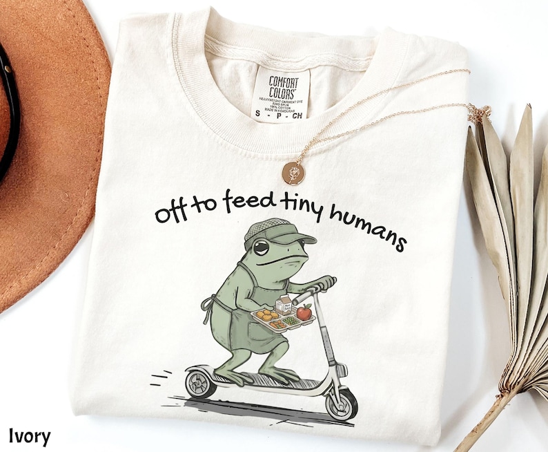 May include: Ivory-colored t-shirt with the phrase "off to feed tiny humans" in black script. The shirt features a cartoon frog wearing an apron and hat, riding a scooter while carrying a tray of food. A gold necklace is draped on the shirt.