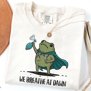 Pode incluir: Uma t-shirt cor de marfim com o gr&aacute;fico de um sapo verde a usar uma capa azul-petr&oacute;leo e um estetosc&oacute;pio, segurando um bal&atilde;o de reanima&ccedil;&atilde;o. Abaixo do sapo, o texto "WE BREATHE AT DAWN" est&aacute; escrito em letras mai&uacute;sculas pretas.
