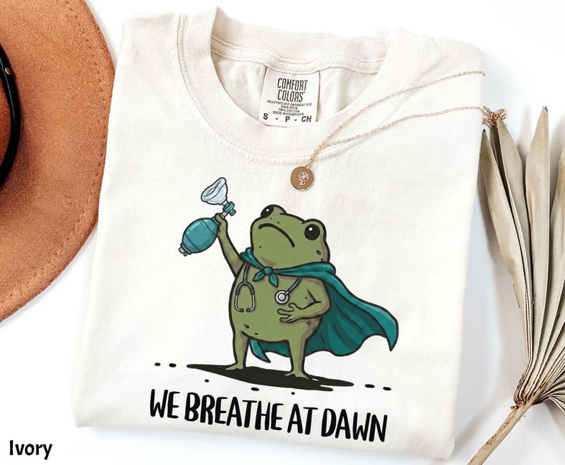 Pode incluir: Uma t-shirt cor de marfim com o gr&aacute;fico de um sapo verde a usar uma capa azul-petr&oacute;leo e um estetosc&oacute;pio, segurando um bal&atilde;o de reanima&ccedil;&atilde;o. Abaixo do sapo, o texto "WE BREATHE AT DAWN" est&aacute; escrito em letras mai&uacute;sculas pretas.