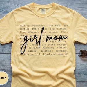 May include: A butter-yellow t-shirt with the words "girl mom" in a handwritten font. The shirt features a list of phrases like "glitter everywhere," "tiny dresses," and "proud girl mama." Sunglasses are on the side.