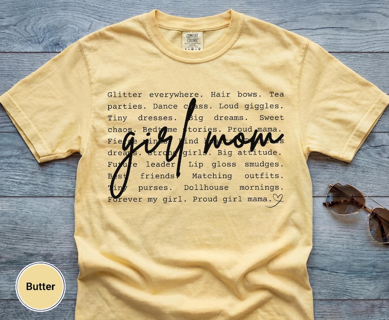 May include: A butter-yellow t-shirt with the words "girl mom" in a handwritten font. The shirt features a list of phrases like "glitter everywhere," "tiny dresses," and "proud girl mama." Sunglasses are on the side.