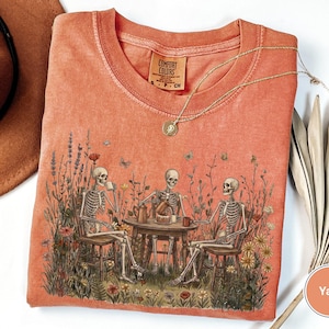 Może przedstawiać: Koralowy t-shirt z nadrukiem graficznym przedstawiającym trzy szkielety siedzące przy okrągłym stole na polu dzikich kwiat&oacute;w, pijące z kubk&oacute;w i jedzące ciasto. Koszulka ma okrągły dekolt i metkę marki Comfort Colors przy szyi.