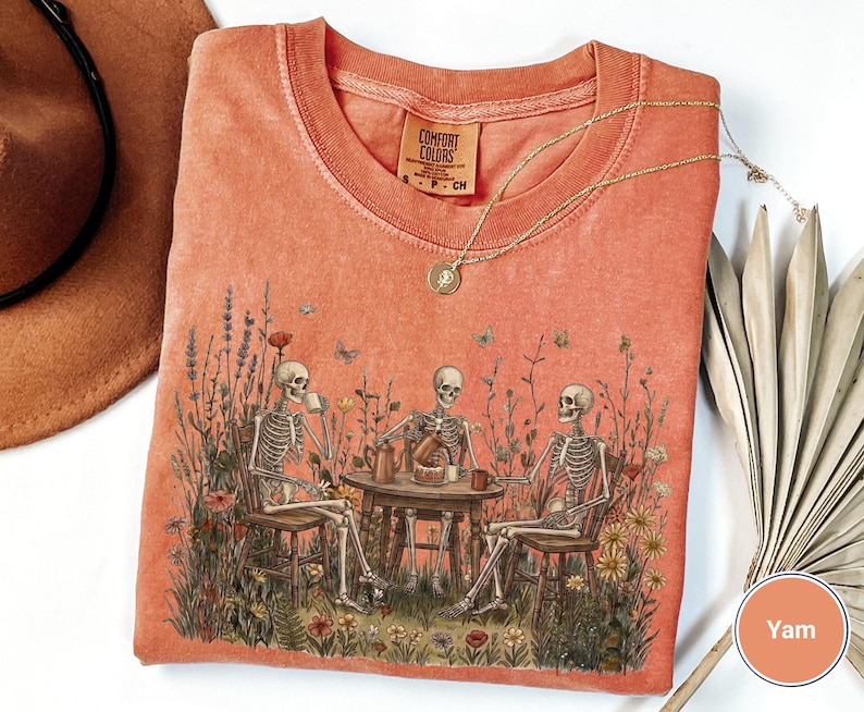 Może przedstawiać: Koralowy t-shirt z nadrukiem graficznym przedstawiającym trzy szkielety siedzące przy okrągłym stole na polu dzikich kwiat&oacute;w, pijące z kubk&oacute;w i jedzące ciasto. Koszulka ma okrągły dekolt i metkę marki Comfort Colors przy szyi.
