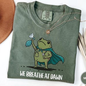Pode incluir: Uma t-shirt de manga curta verde musgo com um gr&aacute;fico de um sapo verde a usar uma capa azul-esverdeada e um estetosc&oacute;pio, segurando um ressuscitador manual. O texto "WE BREATHE AT DAWN" est&aacute; impresso a branco por baixo do sapo.