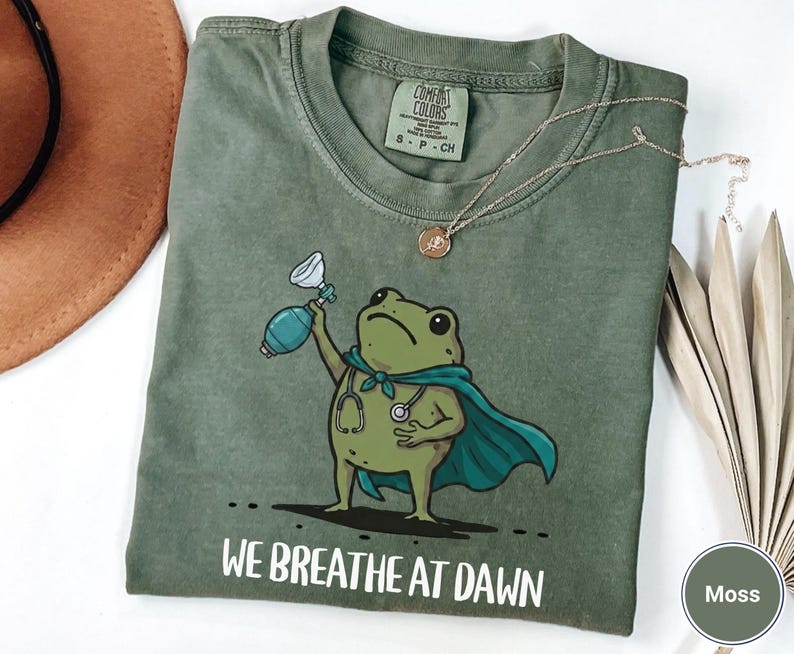 Pode incluir: Uma t-shirt de manga curta verde musgo com um gr&aacute;fico de um sapo verde a usar uma capa azul-esverdeada e um estetosc&oacute;pio, segurando um ressuscitador manual. O texto "WE BREATHE AT DAWN" est&aacute; impresso a branco por baixo do sapo.