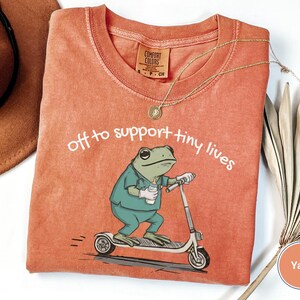 May include: A coral-colored t-shirt with the text "off to support tiny lives." The shirt features a cartoon frog riding a scooter and holding a bottle. A gold necklace and a brown hat are also visible.
