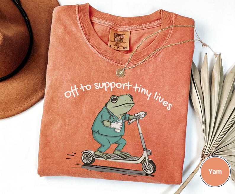 May include: A coral-colored t-shirt with the text "off to support tiny lives." The shirt features a cartoon frog riding a scooter and holding a bottle. A gold necklace and a brown hat are also visible.