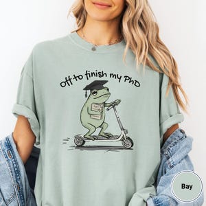 May include: Sage green t-shirt with the phrase "off to finish my PhD" above a cartoon frog riding a scooter. The frog wears a graduation cap and carries books labeled "Draft, Research, and Seminar."