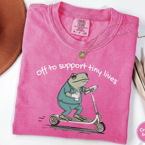 May include: A pink Comfort Colors t-shirt with the text "off to support tiny lives" and a cartoon frog wearing scrubs and riding a scooter. The frog is holding a baby bottle. A gold necklace and a brown hat are also visible.