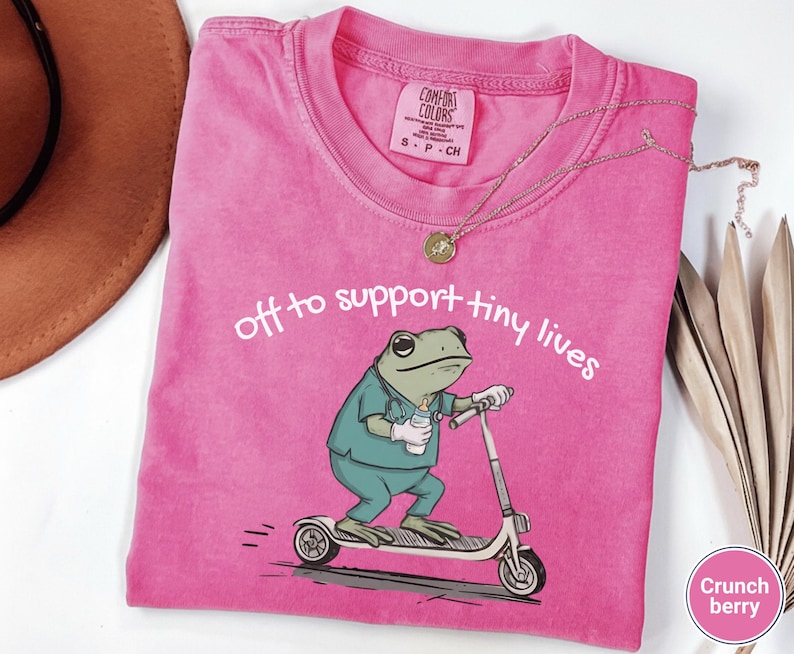 May include: A pink Comfort Colors t-shirt with the text "off to support tiny lives" and a cartoon frog wearing scrubs and riding a scooter. The frog is holding a baby bottle. A gold necklace and a brown hat are also visible.
